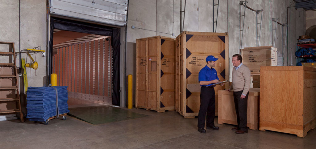 Two men standing in a warehouse with crate behind them.