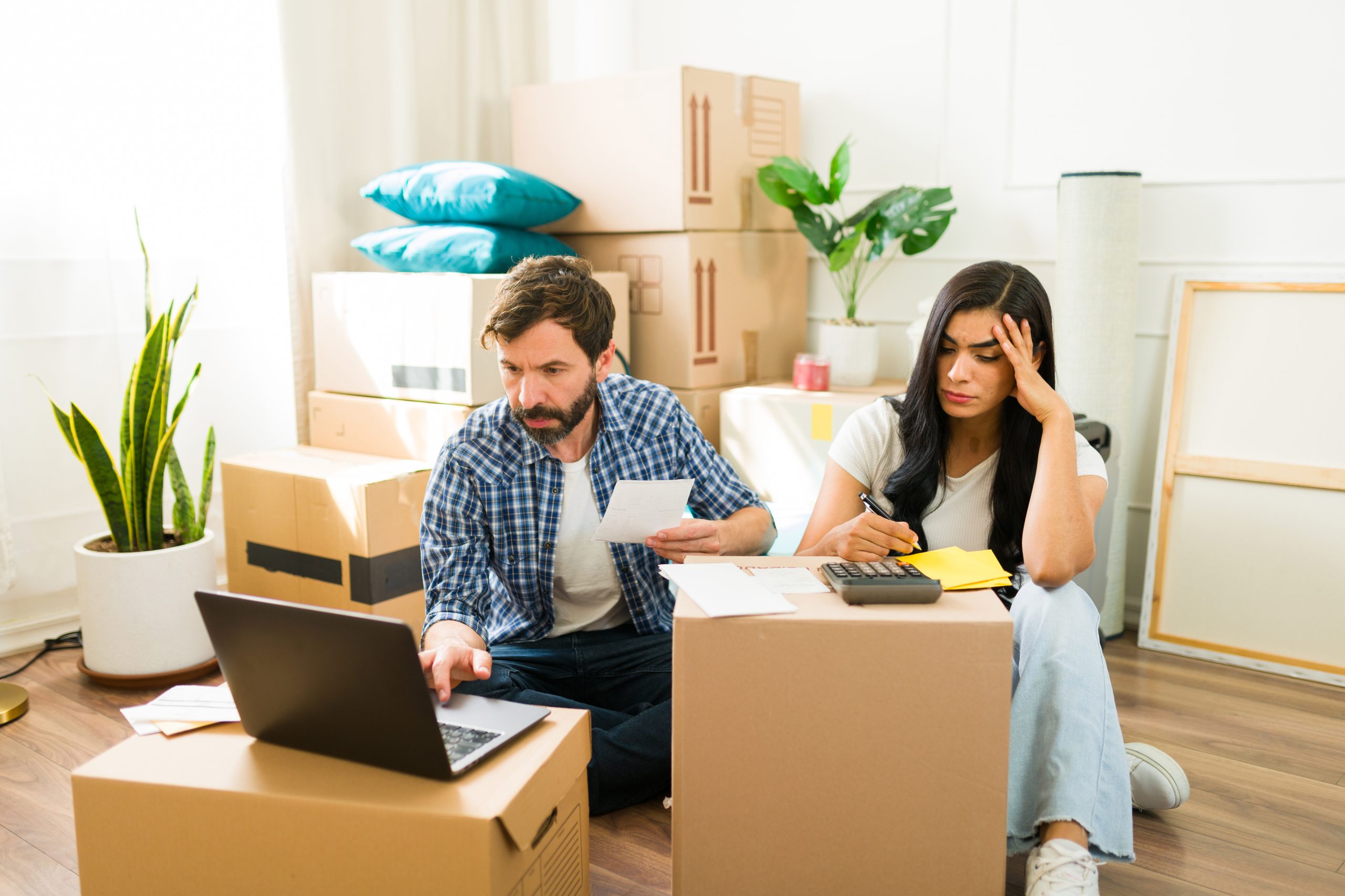 Young couple sitting on the floor surrounded by cardboard boxes and feeling overwhelmed while calculating their moving expenses